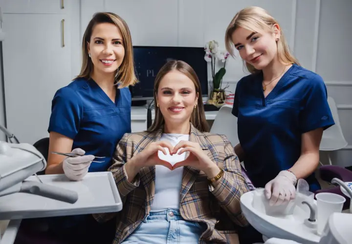 Uśmiechnięta pacjentka tworzy serce dłońmi, otoczona przez dwie dentystki w gabinecie stomatologicznym.
