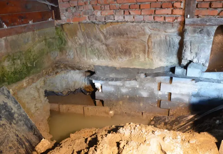 Prace nad hydroizolacją piwnicy od wewnątrz. Widoczny wykop, wilgotne ściany z cegły i kamienia, woda gruntowa.