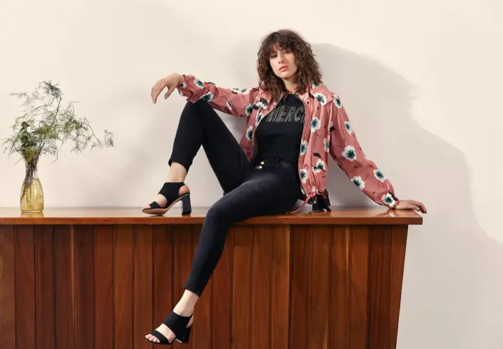 Mujer con rizos sentada en mueble de madera, luciendo distintos tipos de estilos de vestir: chaqueta floral, camiseta con brillos y jeans negros.
