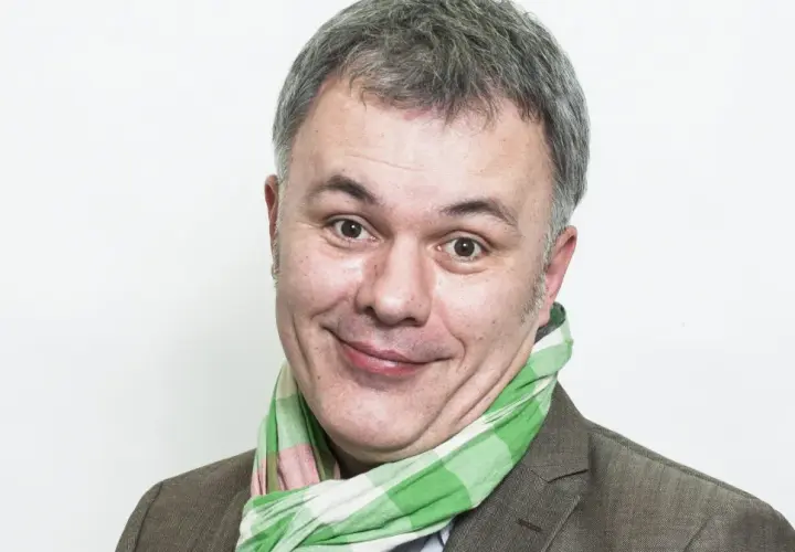 Uśmiechnięty mężczyzna w kratkowanym szaliku, znany jako Mazurek z RMF FM, z szarymi włosami i brązową marynarką.