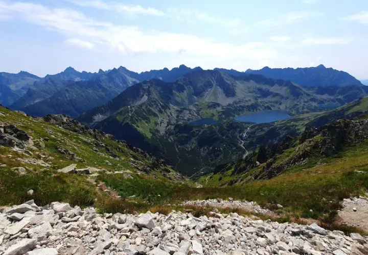 Widok na Tatry z kamienistego zbocza, z jeziorami w dolinie. Szlak turystyczny prowadzi przez malownicze **krzyżne** krajobrazy.