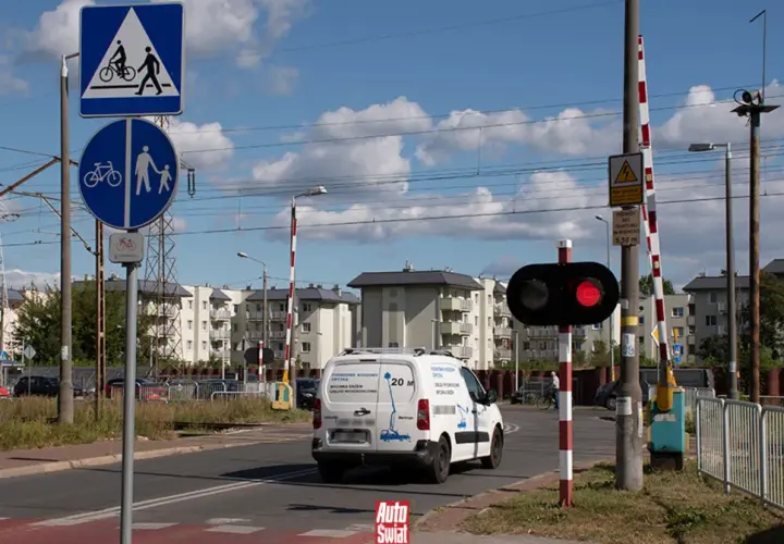 Czerwone światła na przejeździe kolejowym sygnalizują zatrzymanie. Znak informuje o przejściu dla pieszych i rowerzystów.