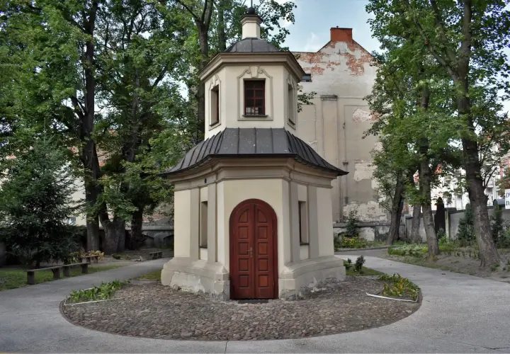 Kapliczka z drewnianymi drzwiami, otoczona drzewami i brukowanym placem. Jedna z urokliwych kaliskich atrakcji.