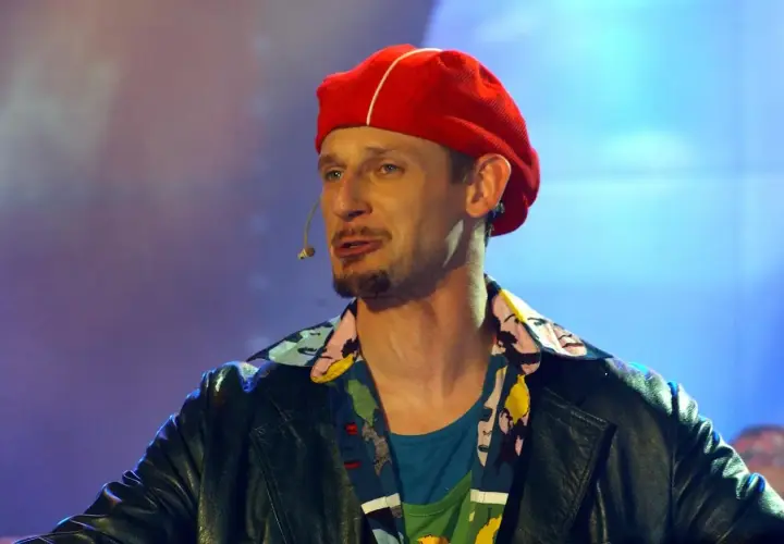 Michał Wójcik w czerwonej beretce i kolorowej koszuli, z mikrofonem przy ustach, na tle niebiesko-fioletowego światła.