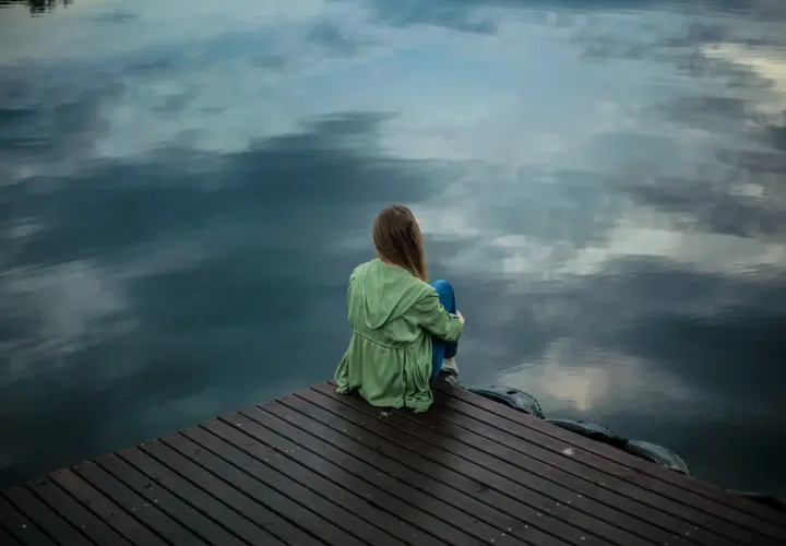 Ein Mädchen sitzt allein auf einem Holzsteg am Wasser. Sie wirkt nachdenklich, als ob sie nicht alleine sein kann.