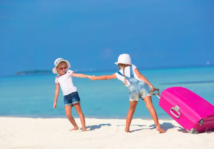 Due bambine si tengono per mano su una spiaggia tropicale, una tira una valigia rosa. Inizia la loro vacanza famiglia mare.