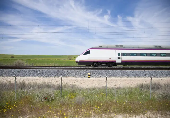Un moderno tren blanco con detalles morados avanza por las vías. El paisaje verde y el cielo azul sugieren un viaje rápido de A Coruña a Madrid.