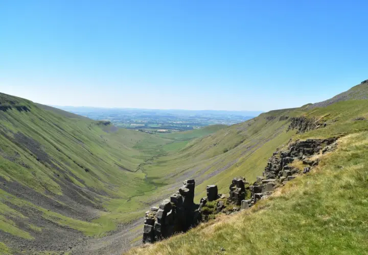 A vast green valley carved by a winding stream, with rocky outcrops in the foreground. This is a classic view along the Pennine Way, under a clear blue sky.