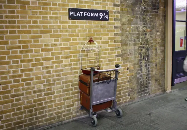 Wózek z bagażami i klatką dla ptaków przy ścianie z napisem "Platform 9 3/4" - magiczne miejsce jak z Kings Cross Harry Potter.