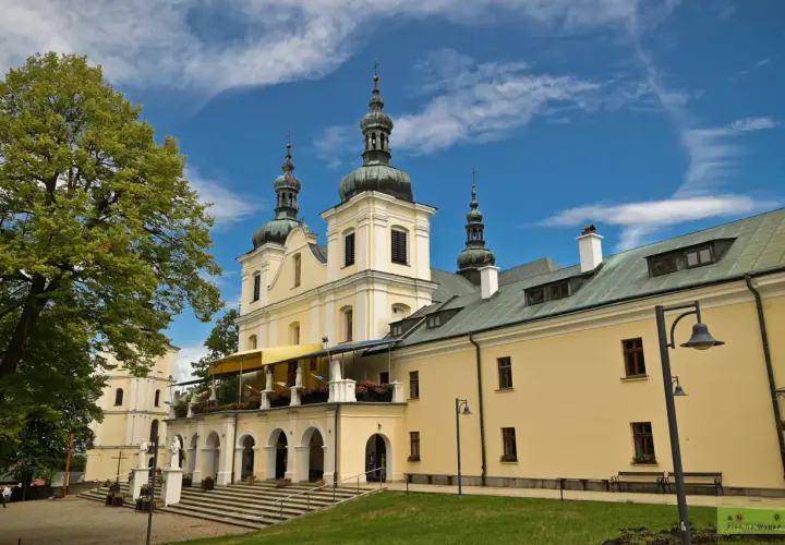 Jasny budynek sanktuarium z kopułami i krzyżami na wieżach, otoczony zielenią. Kalwaria Pacławska dróżki zapraszają do modlitwy.