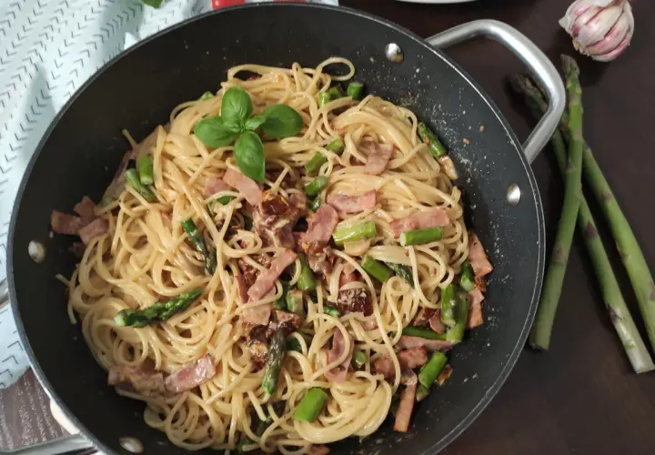 Pyszne spaghetti ze szparagami i boczkiem, udekorowane listkiem bazylii, czeka w patelni. Obok leżą świeże szparagi i czosnek.