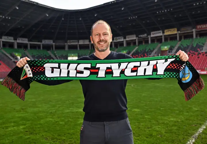 Uśmiechnięty mężczyzna, nowy trener GKS Tychy, trzyma szalik klubowy na tle stadionu.