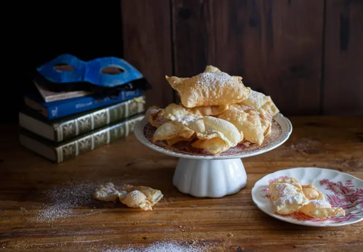 Chiacchiere friabili e bollose, cosparse di zucchero a velo, su un piatto con una maschera veneziana e libri antichi sullo sfondo.
