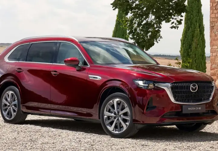 Lśniąca, czerwona Mazda CX-80 zaparkowana na żwirowej drodze, otoczona zielenią i ceglanym murem.