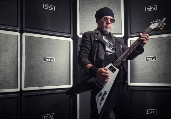 Mężczyzna w czapce i okularach gra na gitarze elektrycznej przed ścianą wzmacniaczy. Wygląda jakby był gotowy na koncert dla fanów popcorn acid drinkers.
