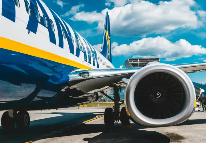 Fragment samolotu Ryanair z widocznym silnikiem i podwoziem na tle błękitnego nieba z chmurami.