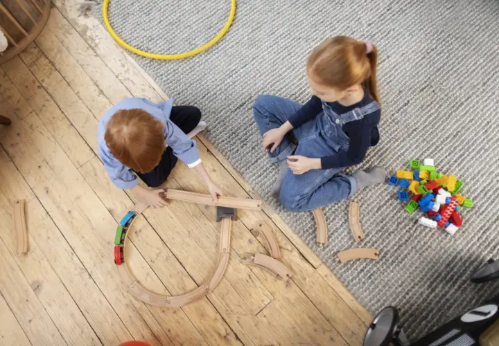 Dzieci budują tory kolejowe, angażując się w odgrywanie ról. Na dywanie leżą klocki, a obok drewniana kolejka.