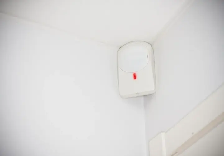 Detector de presencia blanco en esquina de pared, con luz roja encendida. Así es como funciona un detector de presencia.