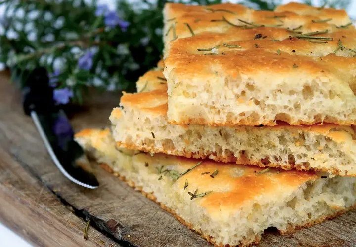 Gestapelte Focaccia-Stücke mit Rosmarin, die an das Backen von Pizza erinnern. Ein Messer liegt daneben auf einem Holzbrett.