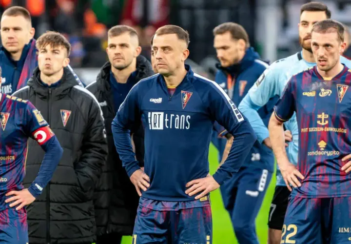 Piłkarze Pogoni Szczecin w ciemnych strojach, z widocznymi herbami klubu. W tle inni zawodnicy i sztab. Wszyscy wyglądają na zamyślonych, być może po trudnym meczu lub w oczekiwaniu na ważne pogoń szczecin - transfery.