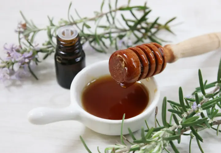 Un vasetto di vetro scuro, un cucchiaio di miele che cola e rametti di rosmarino fresco: ingredienti per un estratto di rosmarino fai da te.