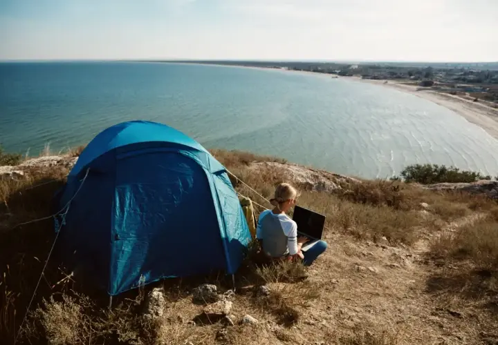 Niebieski namiot i osoba z laptopem na wzgórzu, z widokiem na spokojne, lazurowe morze. Idealny camping lazurowe wybrzeże.