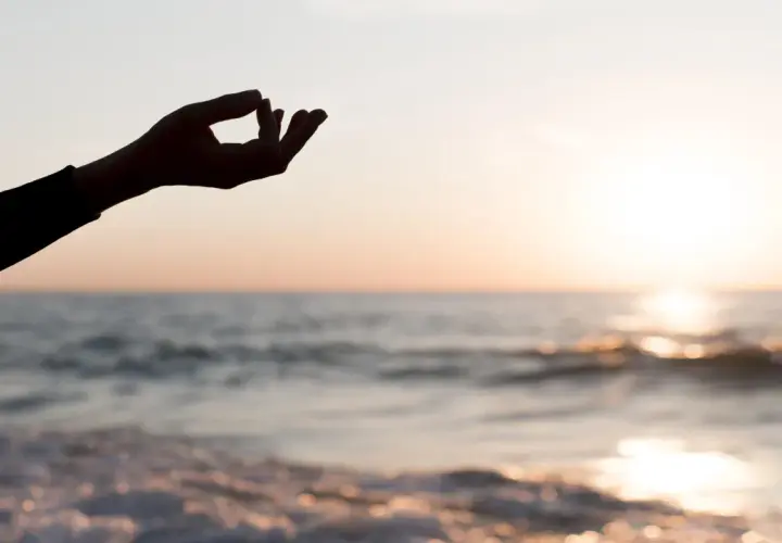Dłoń w geście mudry na tle zachodzącego słońca nad morzem. Chwila głębokiej mindfulness i spokoju.