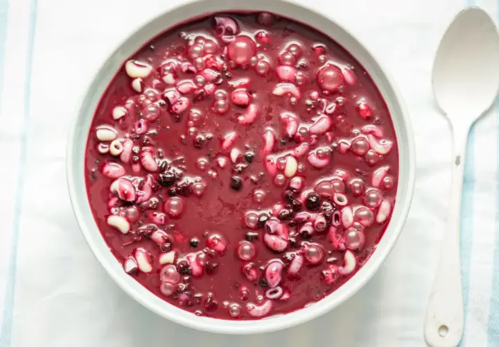 Gęsta, purpurowa zupa jagodowa z makaronem, podana w białej misce obok drewnianej łyżki.