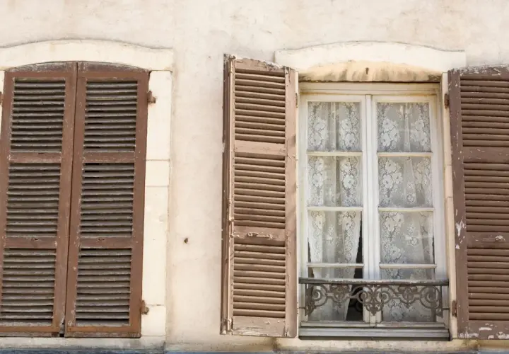 Due finestre d'epoca con persiane in legno marrone. Una è chiusa, l'altra è aperta rivelando tende di pizzo bianco.