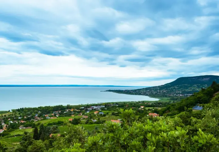 Widok na jezioro Balaton z zielonymi wzgórzami i domkami. Chmury na niebie tworzą dramatyczny krajobraz.