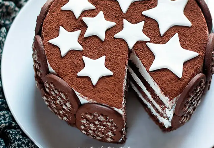 Torta Pan di Stelle ricoperta di cacao, decorata con stelle bianche e biscotti. Una fetta mostra gli strati di crema.