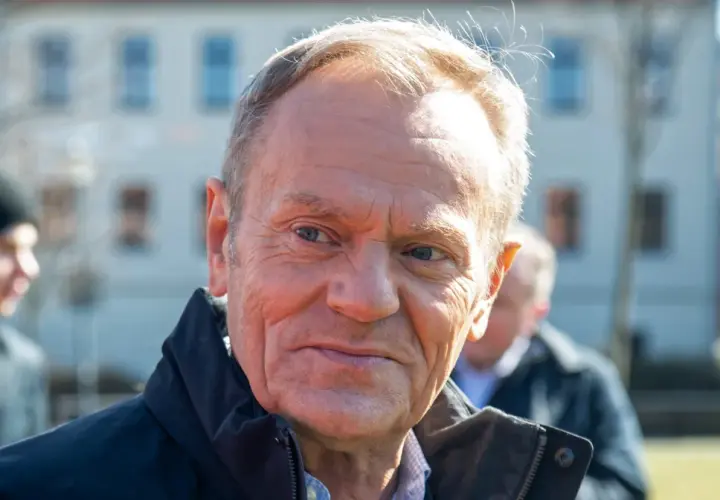 Portret Donalda Tuska w czarnej kurtce, z innymi osobami w tle.