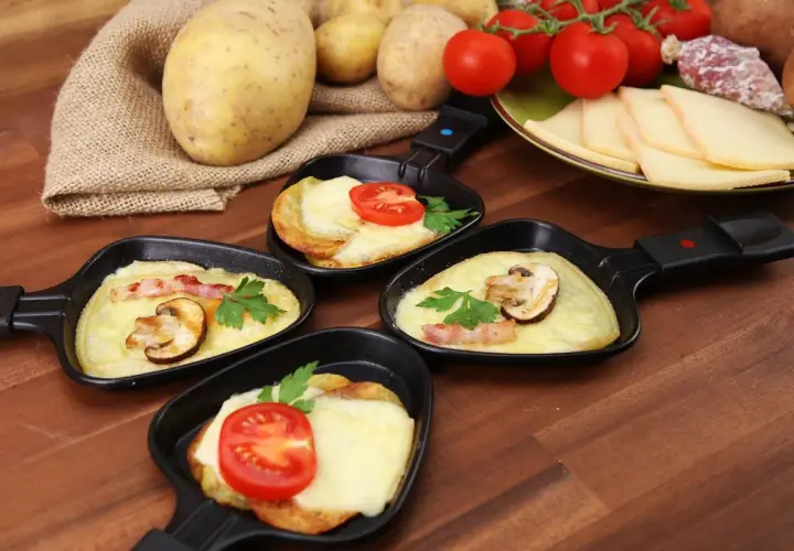 Petites poêles de raclette prêtes à être dégustées, avec pommes de terre, fromage fondu, tomates et champignons. Idéal pour savoir comment couper la raclette.