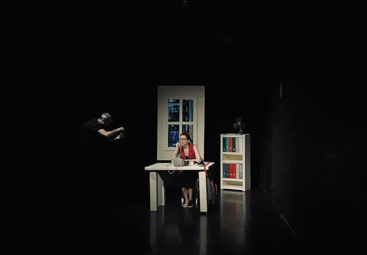 Scena w **teatrze Kana Szczecin**: kobieta przy biurku rozmawia przez telefon, mężczyzna w tle filmuje.