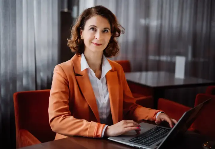 Kobieta w pomarańczowej marynarce pracuje na laptopie w eleganckim wnętrzu.