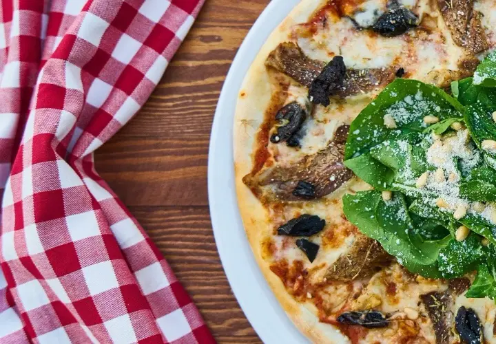 Pizza z rukolą, orzechami pinii i grzybami. Widać kolejność składników na pizzy, od sosu po świeże dodatki.