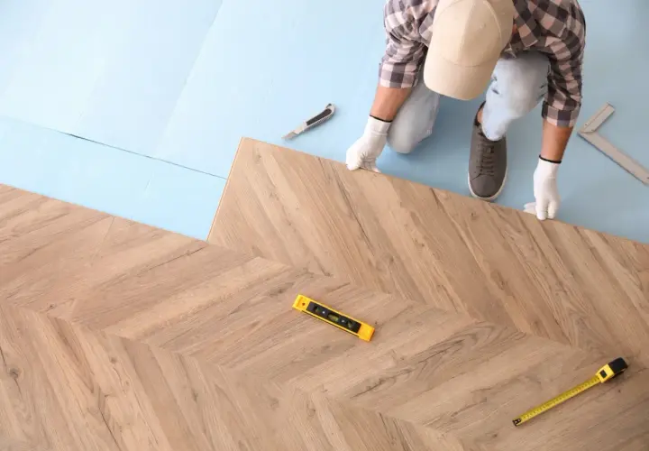 Instalator w białych rękawiczkach układa panele podłogowe w jodełkę. Widać poziomicę i miarkę, co sugeruje, jak kłaść panele.