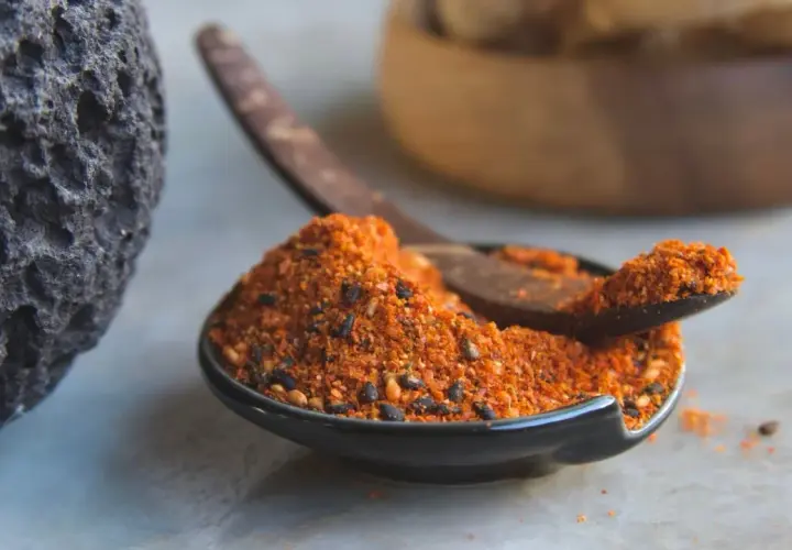 A dark, textured rock sits beside a small bowl filled with a vibrant 7 spice recipe blend. A wooden spoon rests in the bowl, ready to scoop the aromatic mixture.