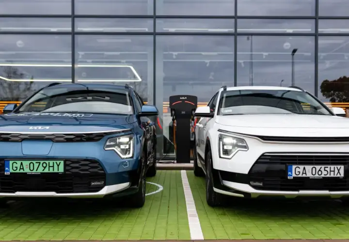 Dwa nowe samochody Kia Niro, jeden niebieski, drugi biały, stoją obok siebie na zielonym parkingu przed nowoczesnym budynkiem z dużymi oknami.