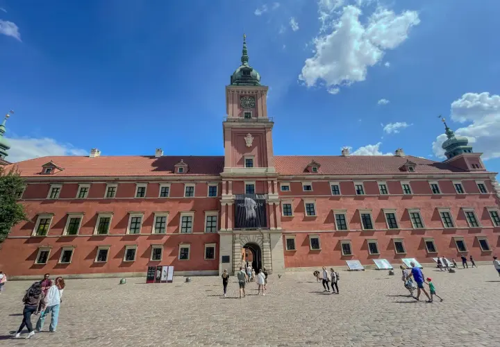 Zamek Królewski w Warszawie, z czerwoną fasadą i wieżą zegarową, na tle błękitnego nieba z chmurami. Plac przed zamkiem tętni życiem.