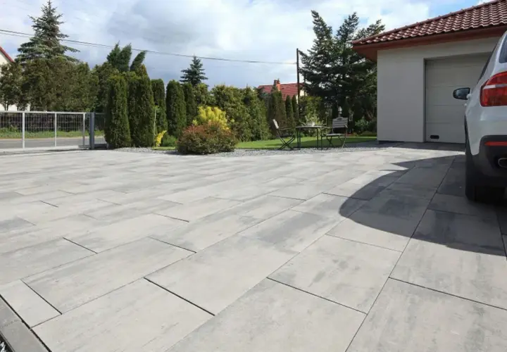 Nowoczesny podjazd z szarej kostki brukowej przed domem z garażem i białym samochodem.