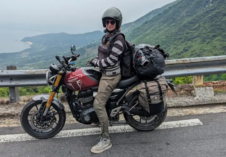 Motociclista con su equipada Triumph Tiger 400, lista para la aventura en una carretera con vistas al mar y montañas.
