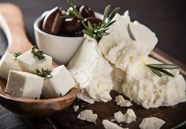Feta, un formaggio ricco di proteine per 100g, servito con olive nere e rametti di rosmarino su un tagliere di legno.