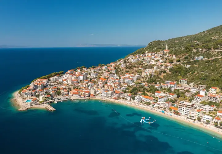 A picturesque village clings to a hillside on the Balkan coast, overlooking a turquoise bay with a sandy beach and inflatable water slides.