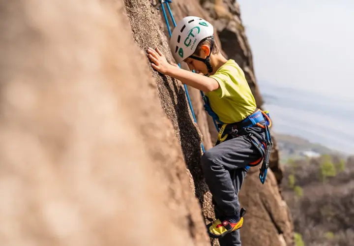 Bambino con casco e imbracatura si arrampica su una parete rocciosa. Un'avventura di arrampicata per bambini, anche se ne avesse 3 anni!