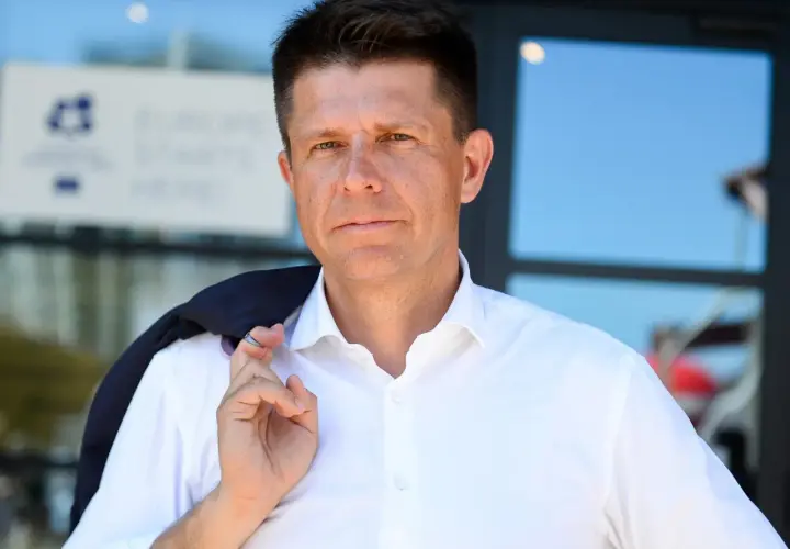 Ryszard Petru w białej koszuli, z marynarką na ramieniu, na tle budynku.