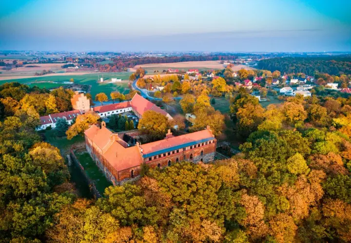 Jesienny krajobraz z zabudowaniami, wśród nich ceglana łubianka otoczona kolorowymi drzewami.