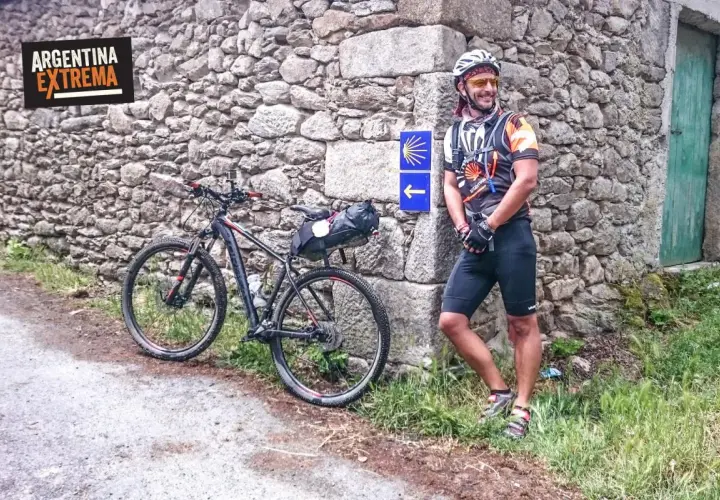 Ciclista sonriente junto a su bici en un camino primitivo, con señal de flecha y concha de peregrino.