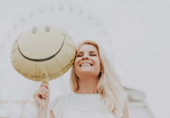 Uśmiechnięta blondynka trzymająca balon w kształcie buźki. Dobre żarty po angielsku poprawiają humor.
