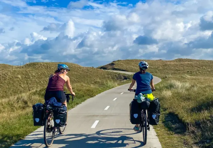 Two cyclists with panniers enjoy a scenic path through grassy dunes, perfect for Netherlands bike trips.